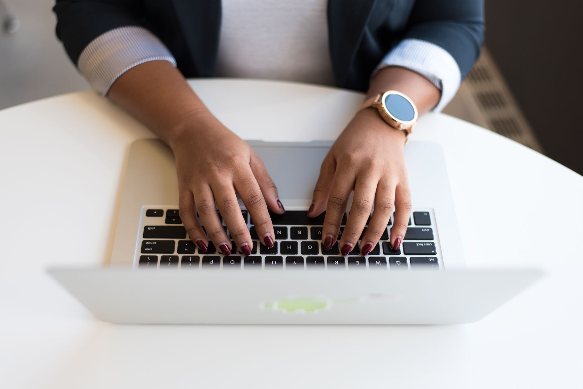 Mains posées sur le clavier d’un ordinateur portable argenté, sur une table blanche, vues du dessus.